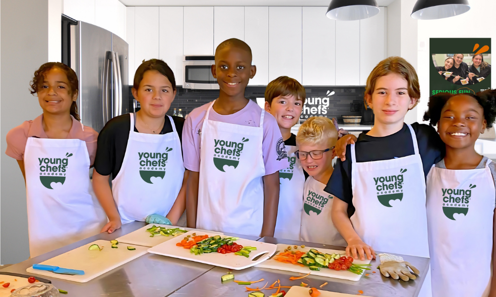 Kids learning to cook at Young Chefs Academy wearing branded aprons in a professional kitchen