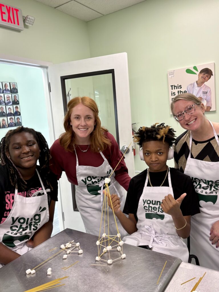 Young Chefs Academy students and instructors smiling around a completed spaghetti tower project in a hands-on cooking and STEM learning class