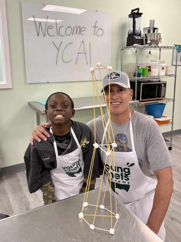Adult and student at Young Chefs Academy posing with a spaghetti and marshmallow tower in a hands-on culinary education class