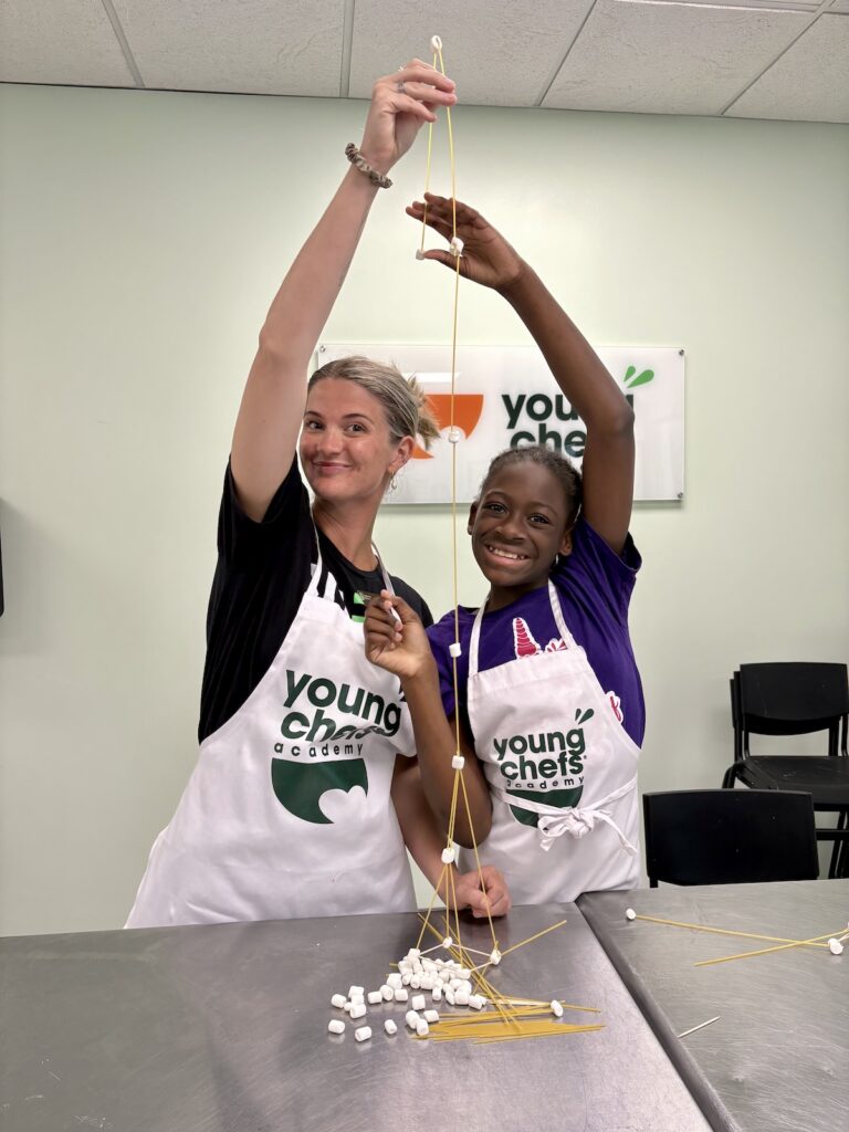 Young Chefs Academy instructor and student building a spaghetti and marshmallow tower during a hands-on STEM cooking class for kids
