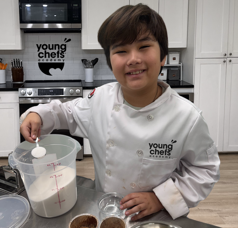 Smiling child in a Young Chefs Academy chef coat measuring ingredients at a kitchen workstation