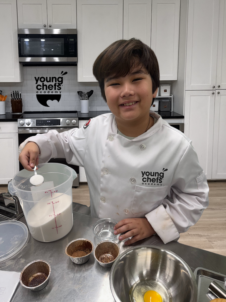 Smiling child in a Young Chefs Academy chef coat measuring ingredients at a kitchen workstation