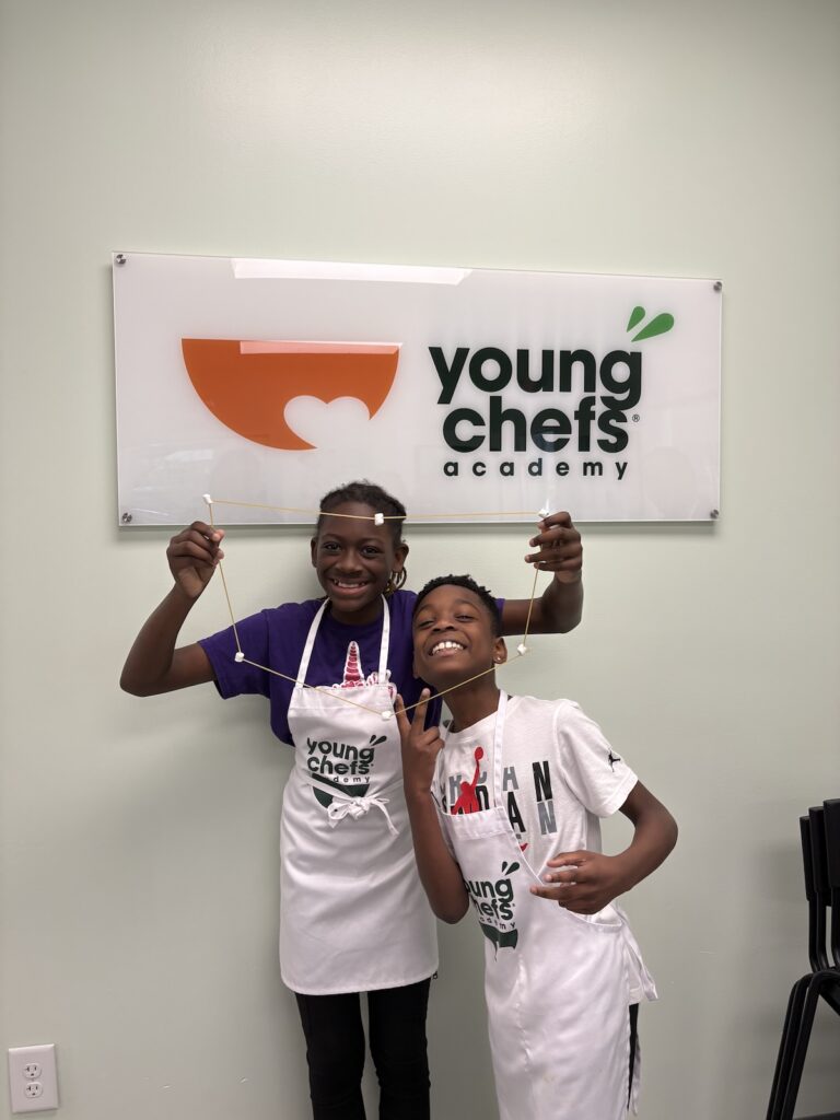 Two students at Young Chefs Academy holding a spaghetti and marshmallow structure during a fun hands-on culinary learning activit